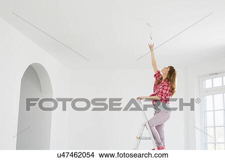 Picture - Full-length of woman on ladder fitting light bulb in new house. Fotosearch
