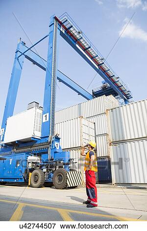 Stock Photo - Full-length of worker using walkie-talkie in shipping yard. Fotosearch