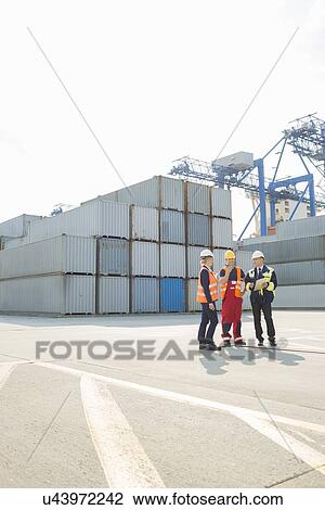 Stock Image - Full-length of workers talking in shipping yard. Fotosearch