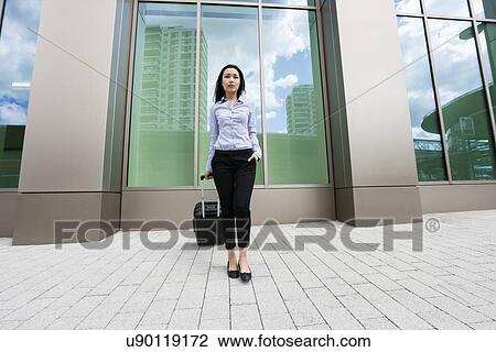 Stock Image - Full length of young businesswoman with suitcase on sidewalk against building. Fotosearch
