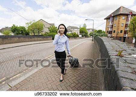 Full length of young businesswoman with luggage while answering cell phone walking on sidewalk View Large Photo Image Stock Image - Full length of young businesswoman with luggage while answering cell phone walking on sidewalk. Fotosearch