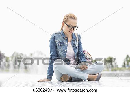 Stock Photo - Full length of young female college student using digital tablet in park. Fotosearch