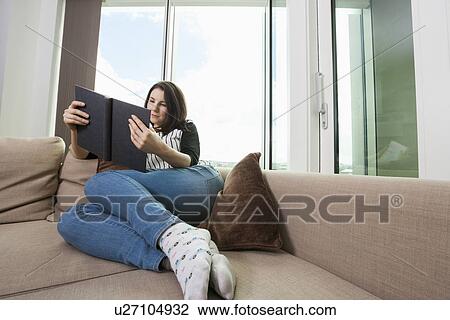 Stock Image - Full length of young woman reading book on sofa. Fotosearch