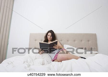 Stock Photo - Full length of young woman reading book in bed. Fotosearch