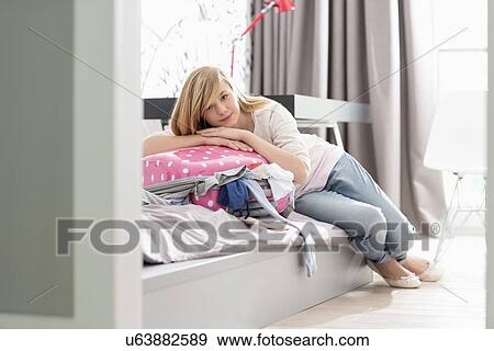 Stock Photo - Full-length portrait of girl relaxing on bulging suitcase at home. Fotosearch