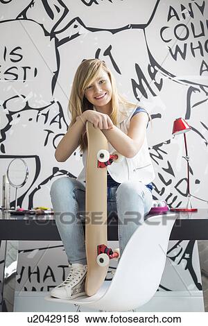 Stock Photo - Full-length portrait of happy teenage girl with skateboard sitting on study table at home. Fotosearch