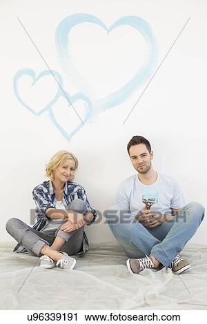 Stock Image - Full-length portrait of mid-adult couple with painted hearts on wall. Fotosearch