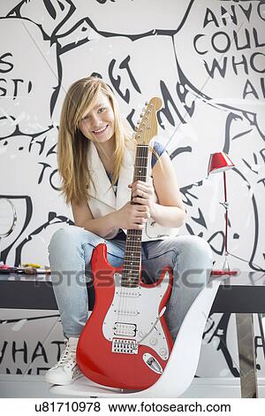 Stock Photo - Full-length portrait of teenage girl with electric guitar sitting on study table at home. Fotosearch