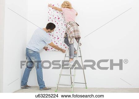 Picture - Full-length rear view of couple applying wallpaper to wall. Fotosearch