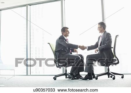 Full-length side view of businessmen shaking hands while sitting on office chairs by window View Large Photo Image Stock Image - Full-length side view of businessmen shaking hands while sitting on office chairs by window. Fotosearch