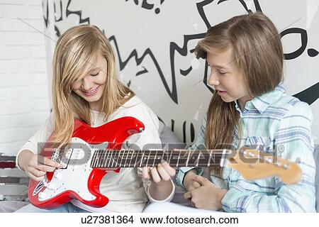 Picture - Girl listening to sister playing electric guitar at home. Fotosearch