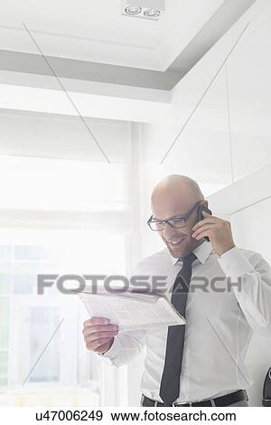 Stock Photo - Happy businessman on call while reading newspaper at home. Fotosearch
