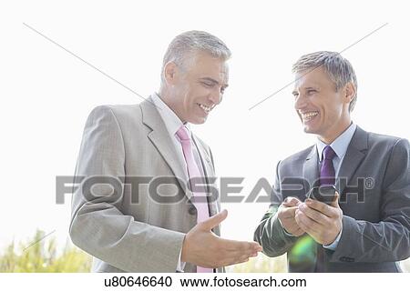 Stock Image - Happy businessmen discussing over mobile phone outdoors. Fotosearch