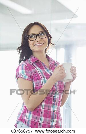 Stock Photo - Happy businesswoman looking away while holding coffee mug in creative office. Fotosearch