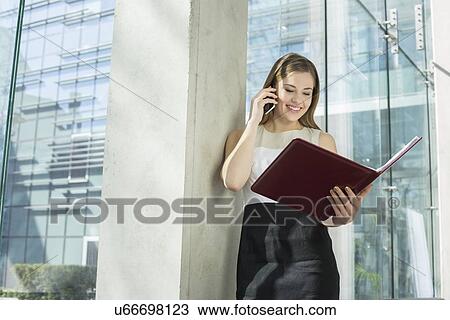 Happy businesswoman using cell phone while reading file in office View Large Photo Image Stock Image - Happy businesswoman using cell phone while reading file in office. Fotosearch