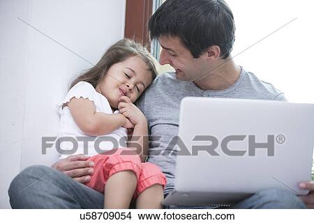 Picture - Happy father looking at cute daughter sleeping while using laptop at home. Fotosearch