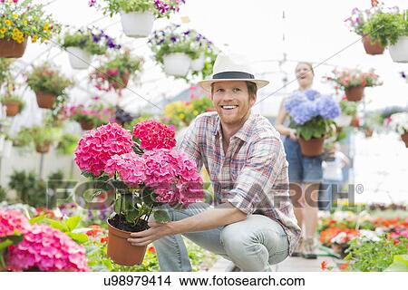 Picture - Happy gardener holding flower pot with colleague standing in background at greenhouse. Fotosearch