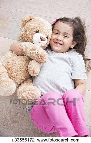 Stock Image - Happy girl with teddy bear lying on wooden floor at home. Fotosearch