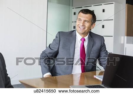 Happy mature businessman sitting at desk in office View Large Photo Image Stock Image - Happy mature businessman sitting at desk in office. Fotosearch