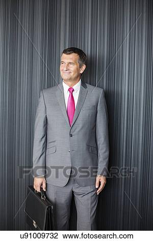 Happy mature businessman with briefcase standing against wall View Large Photo Image Stock Image - Happy mature businessman with briefcase standing against wall. Fotosearch
