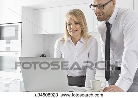 Stock Image - Happy mid adult business couple using laptop at kitchen counter. Fotosearch