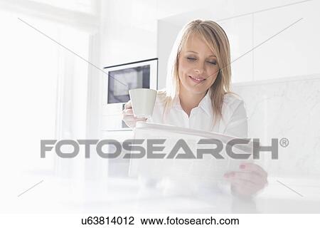 Stock Image - Happy mid adult businesswoman having coffee while reading newspaper at home. Fotosearch
