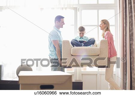 Stock Photograph - Happy parents carrying son on armchair in new house. Fotosearch