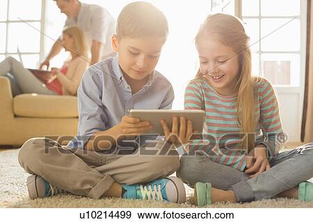 Stock Photo - Happy siblings using digital tablet on floor with parents in background. Fotosearch