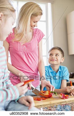 Stock Photo - Happy woman chopping red bell pepper while standing with children in kitchen. Fotosearch