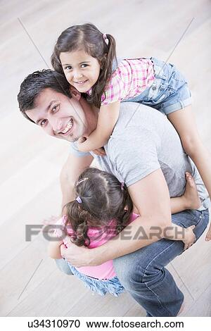 Stock Image - High angle portrait of playful father carrying girls at home. Fotosearch