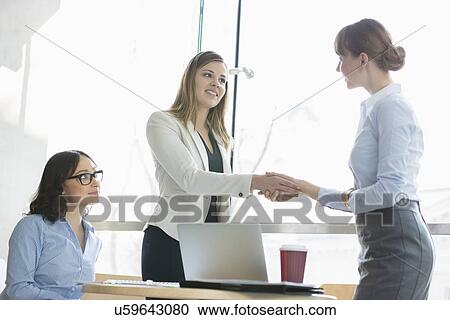 Stock Image - High angle view of businesswomen shaking hands at table in office. Fotosearch