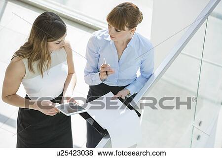 Stock Photo - High angle view of businesswomen discussing over tablet PC and documents by railing in office. Fotosearch