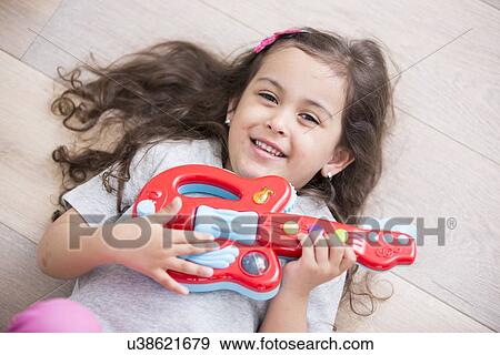 Stock Photo - High angle view of happy girl playing guitar while lying on floor at home. Fotosearch