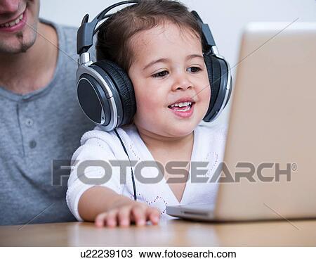 Stock Image - Little girl wearing headphones while looking at laptop with father at table in house. Fotosearch