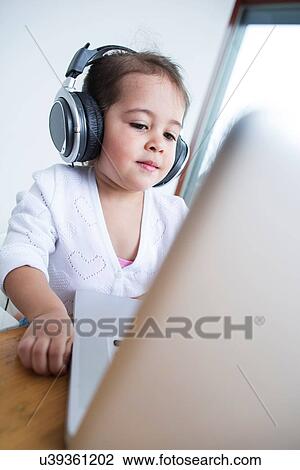 Stock Image - Little girl wearing headphones while looking at laptop at table in house. Fotosearch