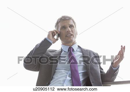 Stock Image - Low angle view of businessman talking on cell phone against sky. Fotosearch