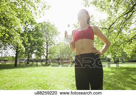 Stock Image - Low angle view of fit woman listening to music while holding water bottle in park. Fotosearch