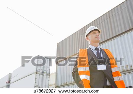 Stock Photo - Low angle view of worker standing against cargo containers in shipping yard. Fotosearch