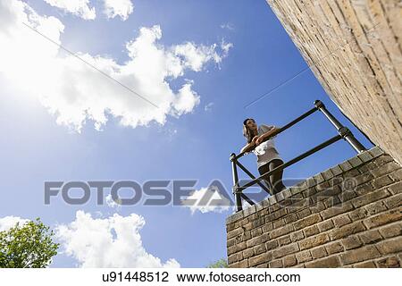 Stock Image - Low angle view of young woman leaning on railing against cloudy sky. Fotosearch