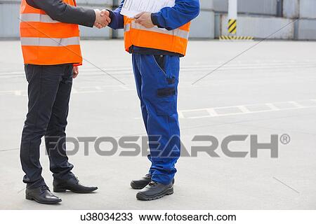 Stock Image - Low section of workers shaking hands in shipping yard. Fotosearch