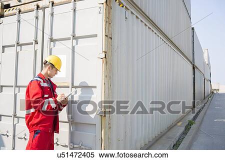 Stock Photography - Male worker inspecting cargo container while writing on clipboard in shipping yard. Fotosearch
