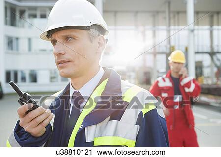 Stock Image - Male worker using walkie-talkie with colleague in background at shipping yard. Fotosearch