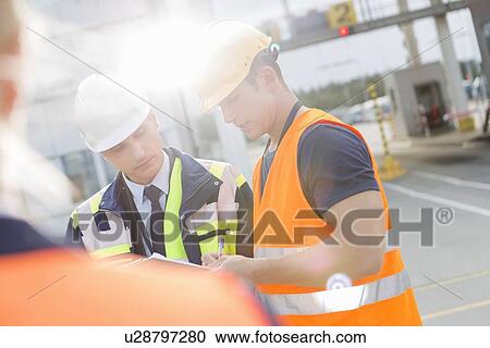 Stock Image - Male workers discussing over clipboard in shipping yard. Fotosearch