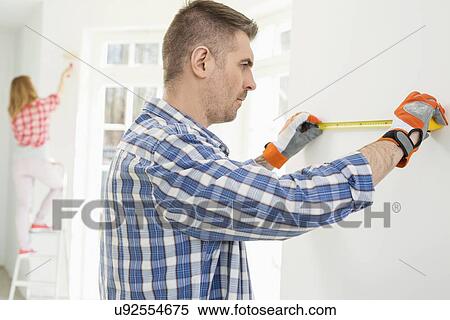 Stock Photography - Man measuring wall with woman painting in background. Fotosearch