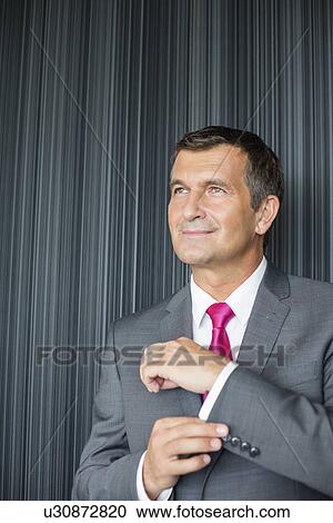Stock Image - Mature businessman buttoning sleeve against wall. Fotosearch