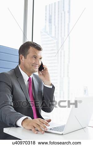 Mature businessman talking on cell phone while using laptop in office View Large Photo Image Stock Photo - Mature businessman talking on cell phone while using laptop in office. Fotosearch