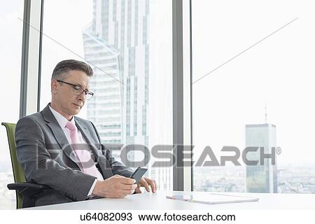 Stock Image - Mature businessman text messaging through cell phone near office window. Fotosearch