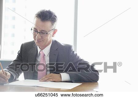 Stock Photography - Mature businessman writing on book at table in office. Fotosearch