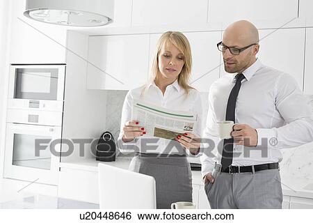 Stock Photograph - Mid adult business couple reading newspaper while having coffee in kitchen. Fotosearch