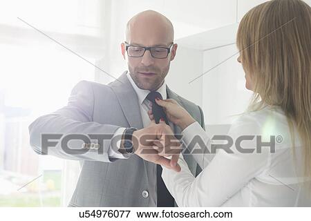 Mid adult businessman checking wristwatch while woman adjusting his tie at home View Large Photo Image Stock Photo - Mid adult businessman checking wristwatch while woman adjusting his tie at home. Fotosearch